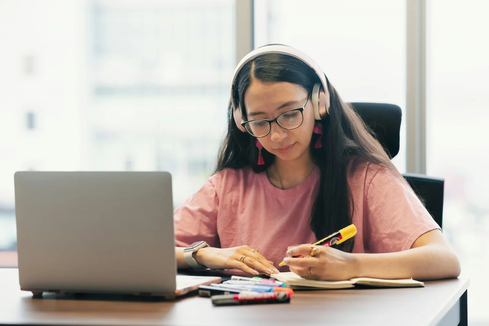 A student wearing headphones, writing in a notebook beside an open laptop during an online lesson.