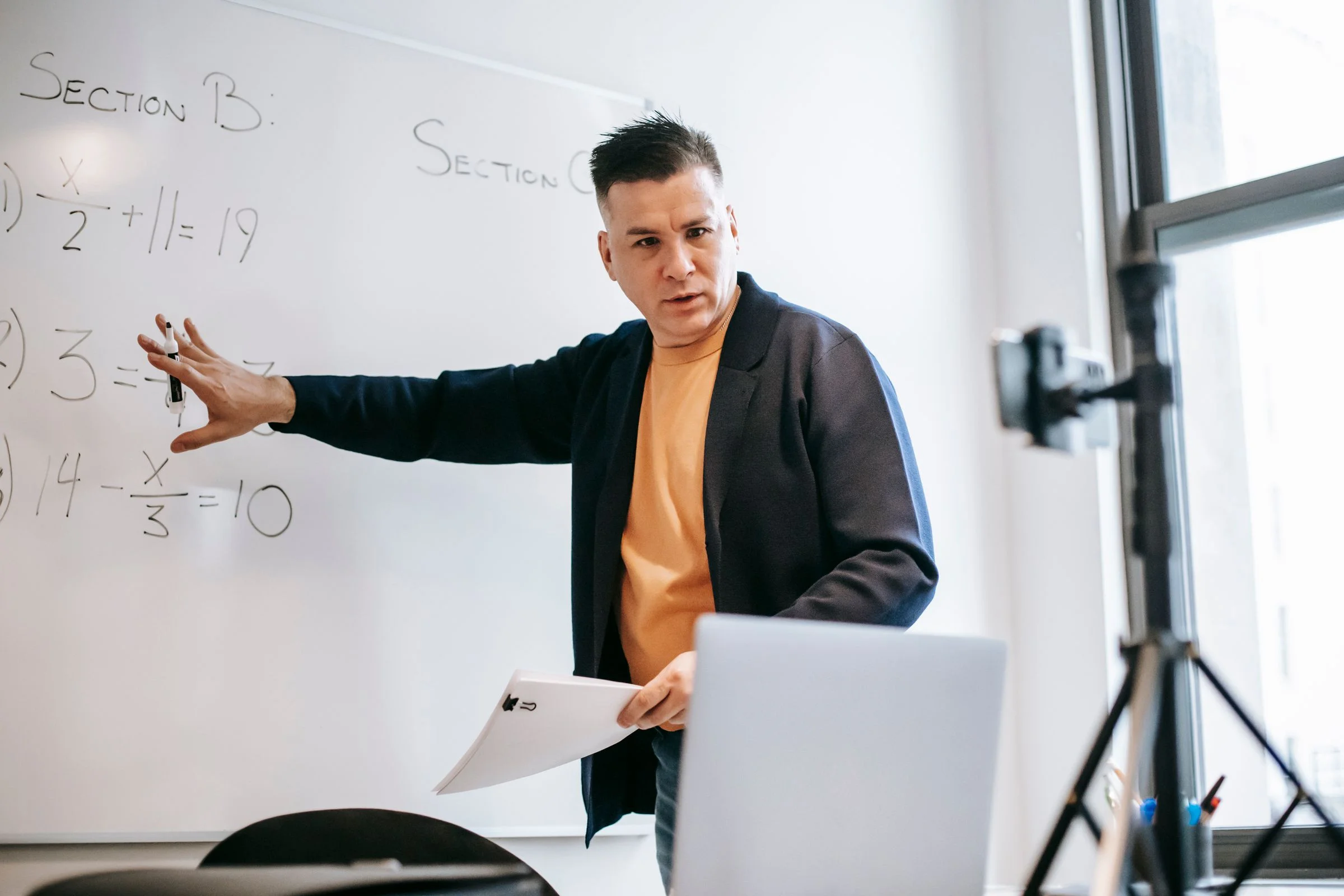 A tutor explaining a step at the whiteboard beside his laptop during an online lesson