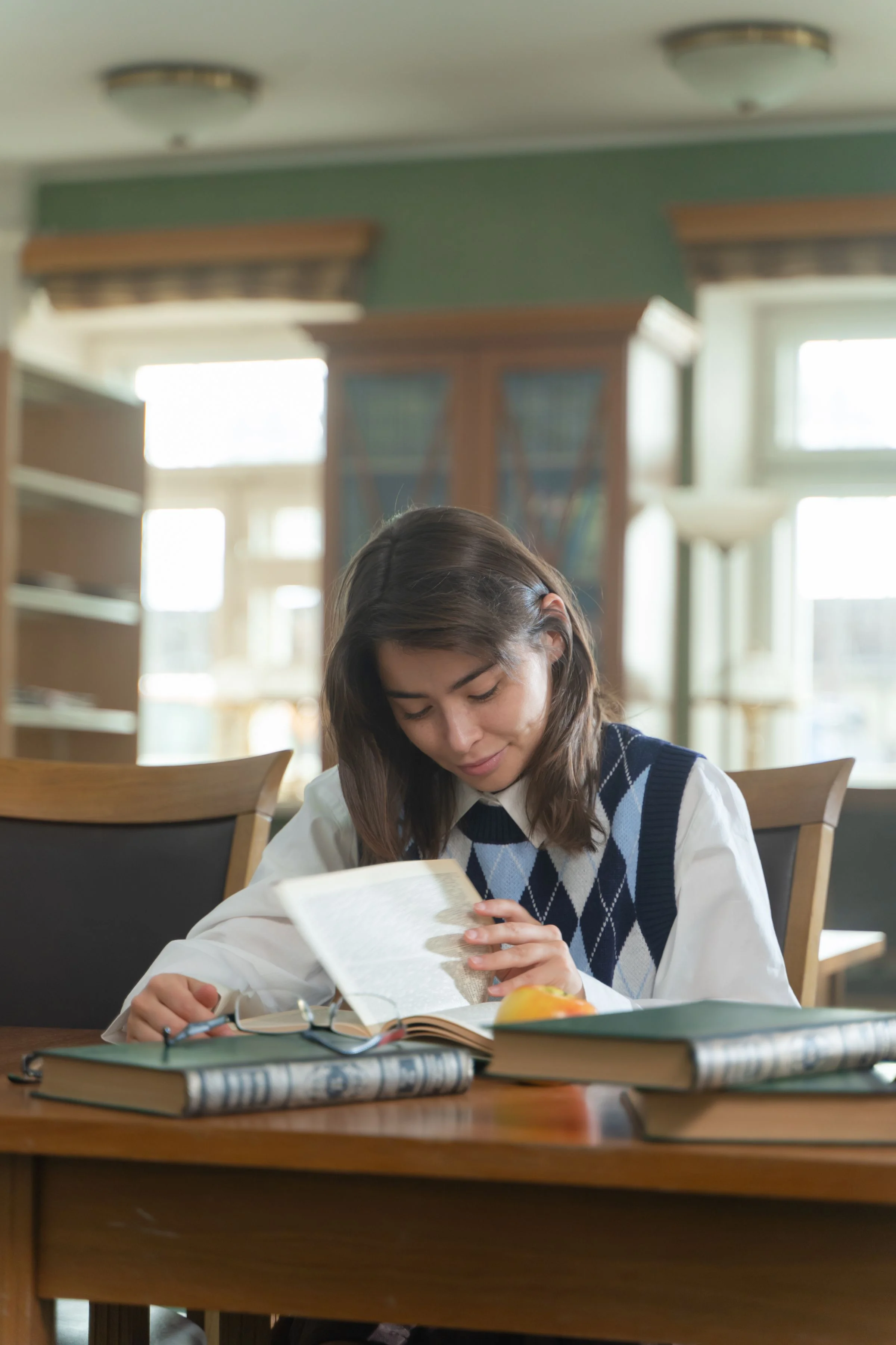 A sixth-form student reading a book by a large window in warm light.